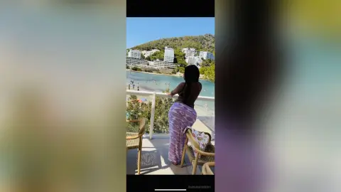 Woman in a purple patterned maxi skirt relaxing on a balcony overlooking a sunny Mediterranean coastal town.