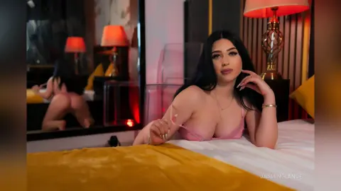 JasminGlance lies on a white bed wearing a pink bra, showing her cleavage and long dark hair in a bedroom setting.