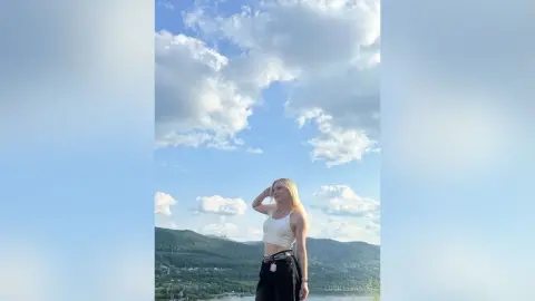 Blonde woman in a white crop top and black pants standing under a bright blue cloudy sky.