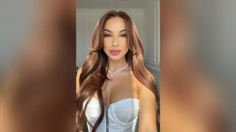 Beautiful woman with long wavy brown hair wearing a white corset and pearl necklace in a bright room.