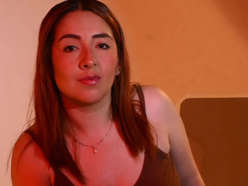 Young woman with long brown hair wearing a brown tank top in warm, moody indoor lighting.