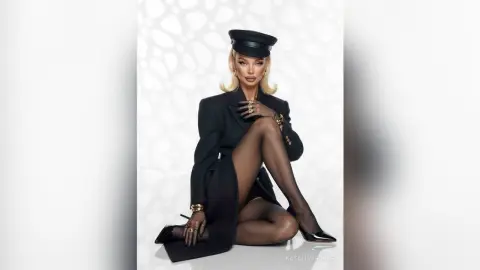 Elegant woman in a black blazer and cap posing glamorously against a white textured background.