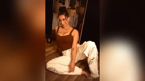 Stylish woman in a brown tank top and ripped white jeans sitting on a wooden floor in a warm indoor setting.
