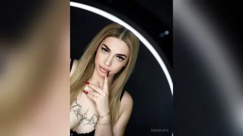 ZaraPlaya poses with a finger to her lips, showing her long blonde hair, red nails, and a large chest tattoo or chest piece.