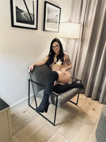IvyRaewants2play sits naked in a grey chair wearing black thigh-high boots and glasses, showing her bare breasts and bare buttocks.