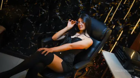 Elegant woman in a black dress and glasses relaxing in a luxury black leather chair against a dark marble wall.