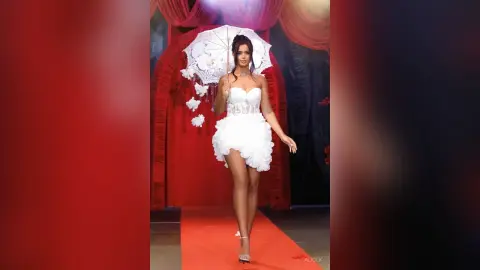 Elegant woman in a white ruffled cocktail dress walking on a red carpet under a white lace parasol.
