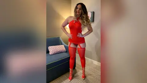 Smiling woman in a red bodysuit and matching thigh-high boots posing in a modern living room.