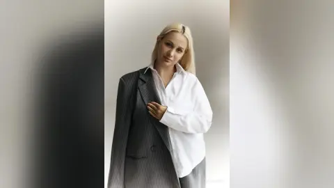 Elegant blonde woman in a white button-down shirt and grey pinstripe blazer posing in a bright minimalist studio.