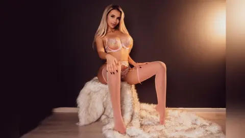 Blonde woman in pink lace lingerie posing on a white faux fur rug in a moody, dimly lit studio setting.