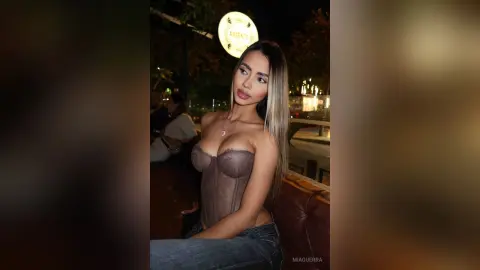 MiaGuerra poses in a brown corset and jeans, showing off her cleavage and long blonde hair while sitting outdoors at night.
