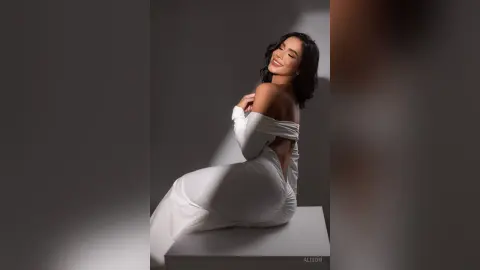 Alison sits on a white platform wearing a white off-the-shoulder dress that shows her bare back and shoulders.