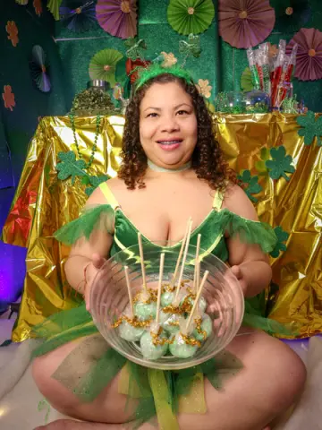 Smiling woman in a green fairy costume holding a bowl of mint cake pops at a festive St. Patrick's Day party.