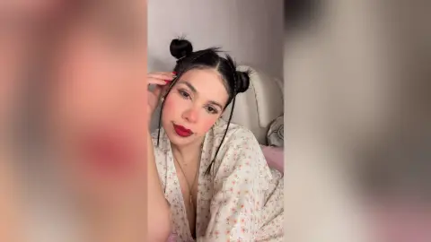 Young woman with space buns and red lipstick wearing a floral robe in a cozy indoor setting.