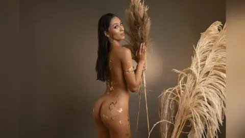 Nude woman with gold body glitter posing elegantly amidst tall pampas grass in a warm studio setting.