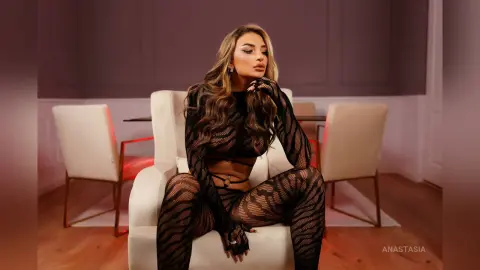 Anastasia sits on a white chair wearing a black mesh long-sleeve top and matching patterned leggings.