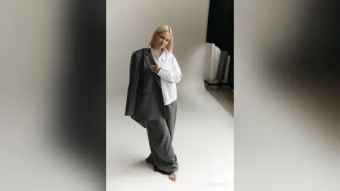 Blonde woman in oversized grey blazer and white shirt posing in a minimalist studio setting.
