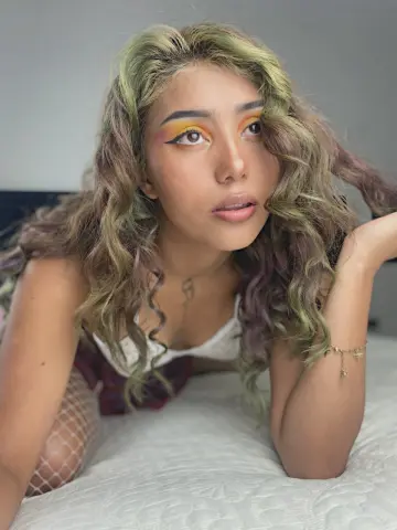 Young woman with wavy green hair and colorful eye makeup wearing a white lace top on a white bed.