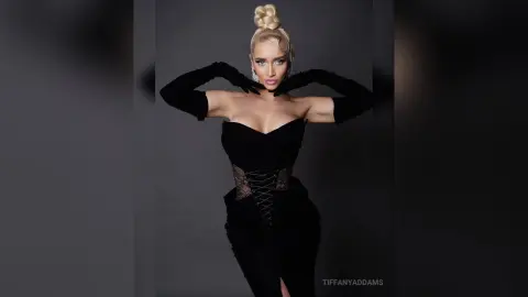 TiffanyAddams poses in a black lace-up corset and long black gloves, showing off her blonde hair tied in a high bun against a dark background.