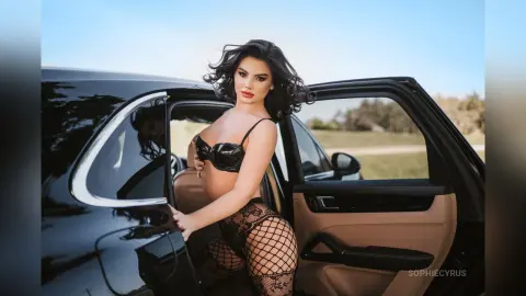 Sofiya poses against a black car, wearing a black bra and fishnet stockings.