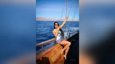 MissyJolie sits on the wooden railing of a boat in a white patterned bikini and sunglasses against the blue ocean.