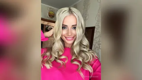Smiling blonde woman in a bright pink blouse posing in a cozy, sunlit room.