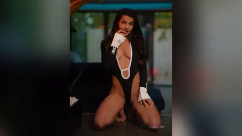Glamorous woman in a black plunging bodysuit posing in a modern, moody indoor setting.