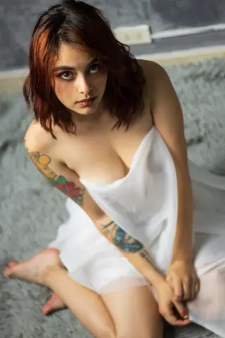 Young woman with reddish hair in a white sheer wrap sitting on a grey shag rug in a moody indoor setting.