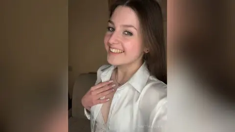 Smiling young woman in a white button-down shirt posing in a cozy indoor setting.