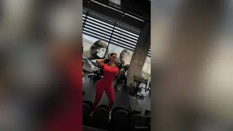 NicoleBella wears a red bodysuit and white headphones while standing in a gym setting near several black dumbbells.