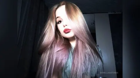 Young woman with long pastel pink hair and red lipstick posing in a moody indoor setting.