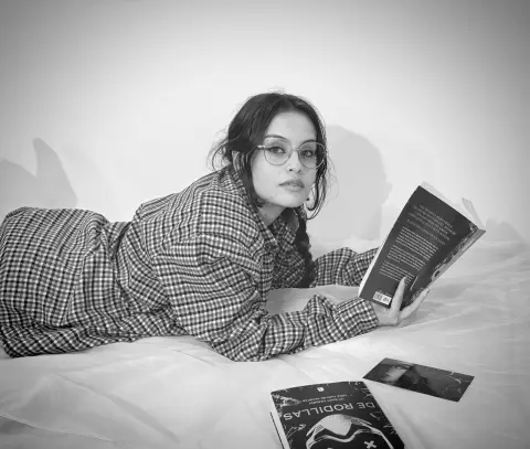 Young woman in checkered pajamas reading a book on a white bed in a moody black and white portrait.
