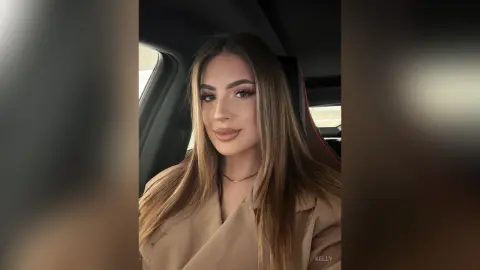 Young woman with long blonde hair wearing a beige trench coat sitting in a car with a soft, natural glow.