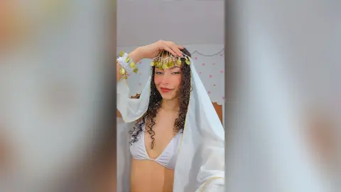 Smiling woman in white bikini and gold coin headpiece posing in a dreamy, ethereal setting.