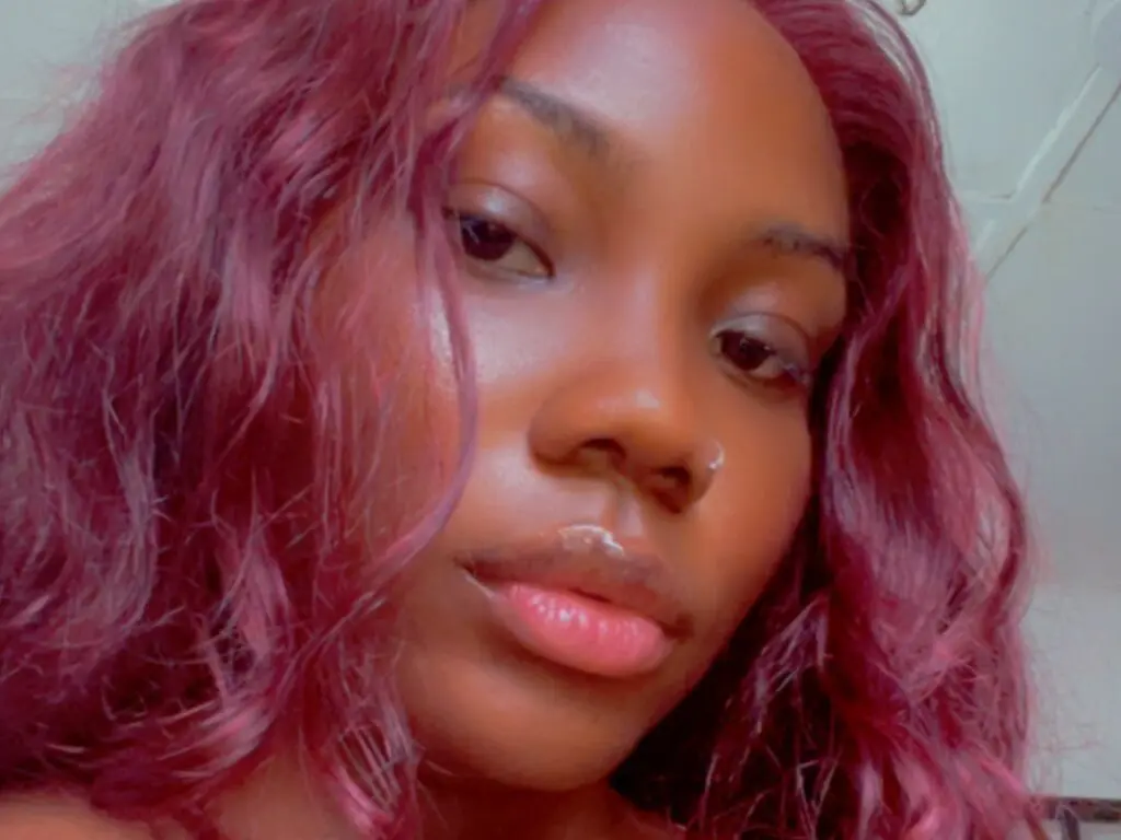 Close-up portrait of a young woman with wavy magenta hair and glowing skin in soft indoor lighting.