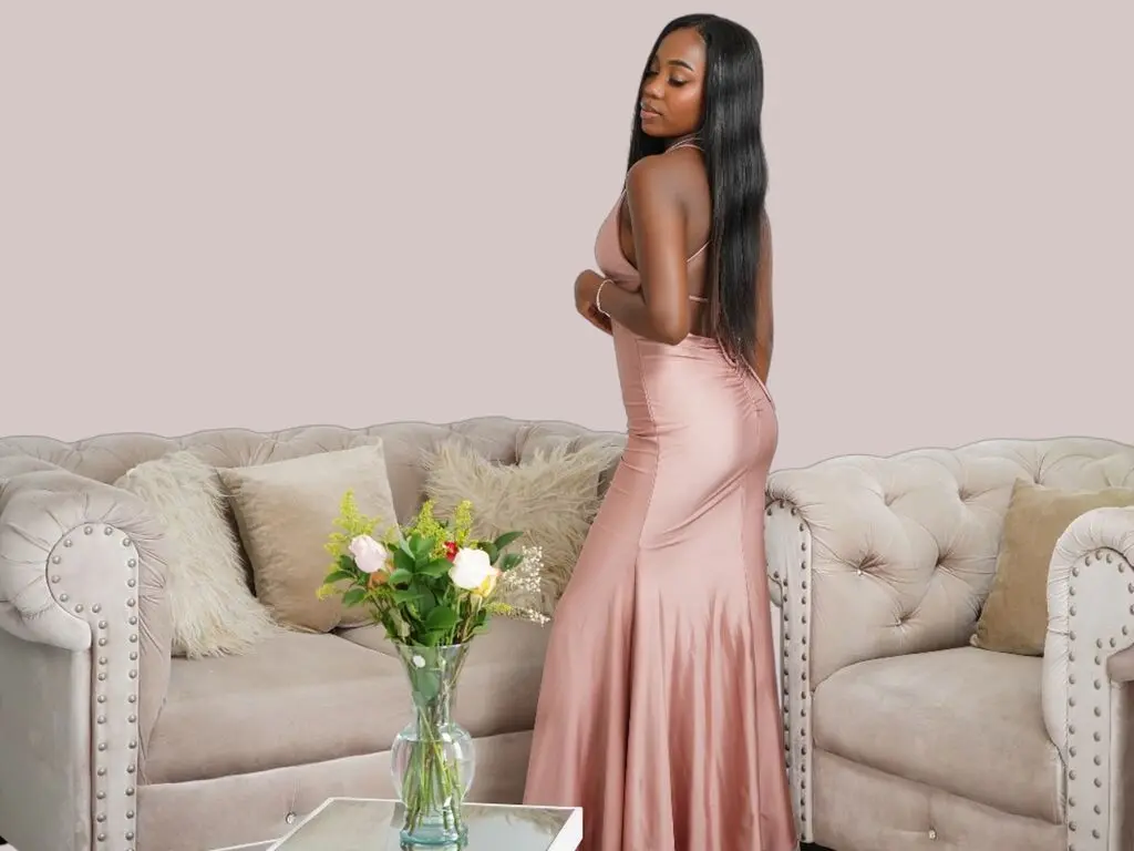 Elegant woman in a rose gold satin evening gown posing in a neutral living room setting.