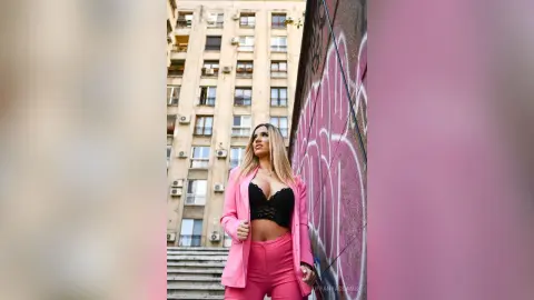 TiffanyAddams poses in a pink blazer and matching pink trousers with a black lace bra visible underneath.