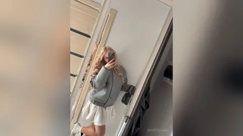 KrystalLaaz takes a mirror selfie wearing a grey cardigan and a short white skirt.