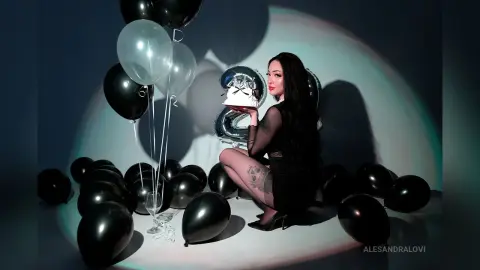 Elegant woman in a black dress sitting among black and silver balloons in a moody studio setting.