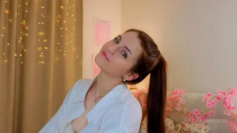 Smiling woman in a white button-down shirt sitting in a cozy room with fairy lights and pink floral decor.