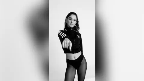 Stylish woman in black Adidas tracksuit and tights posing confidently in a minimalist black and white studio setting.