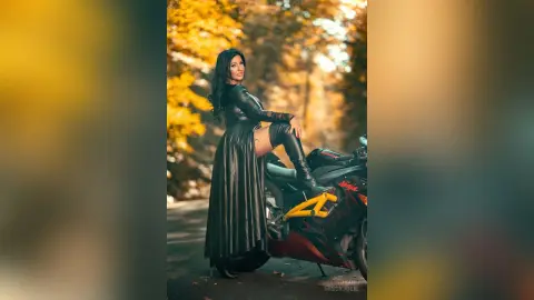 MissyJolie poses next to a black motorcycle wearing a black leather top and a long, slit-leg skirt or skirt-trousers combo.