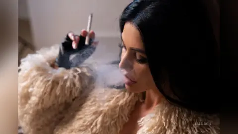 Moody portrait of a brunette woman in a fuzzy cream coat smoking a cigarette in a dimly lit setting.