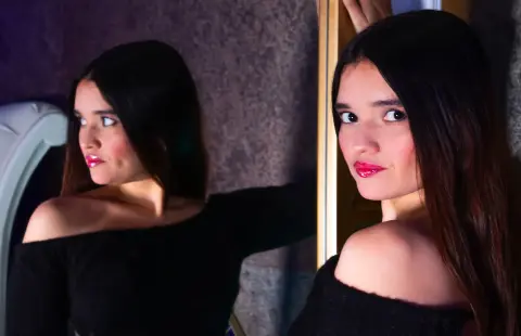 Young woman with long dark hair in a black off-the-shoulder top posing in a moody, dimly lit room.