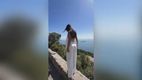 IsabellaMatters stands overlooking the ocean, wearing white high-waisted trousers and showing her long dark hair and bare back.