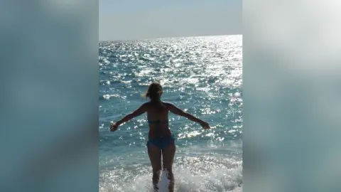 Young woman in a blue bikini standing on a sunny beach facing the sparkling ocean waves.