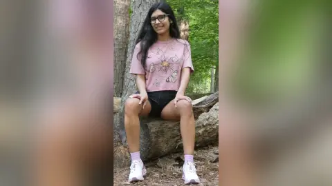 Smiling young woman in a pink butterfly graphic t-shirt and black shorts sitting on a log in a lush green forest.