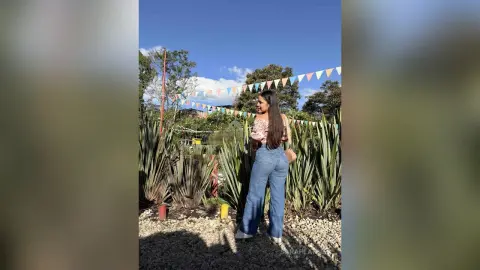 Young woman in high-waisted blue jeans and a floral blouse standing in a sunny garden with colorful bunting.