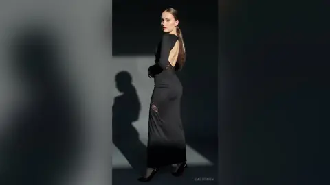 EmilyGiffin poses in a long black dress with a low open back and lace cutouts, standing against a dark wall with a shadow behind her.
