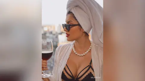 Stylish woman in a white robe and pearls holding a glass of red wine on a city balcony at sunset.